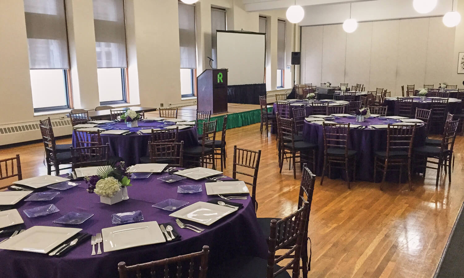 Large room with multiple dining tables set up for reception, with a speaker podium and presentation screen.