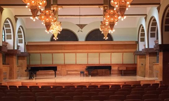 Indoor view of Ganz Hall showcasing a stage with piano and theater style seating.