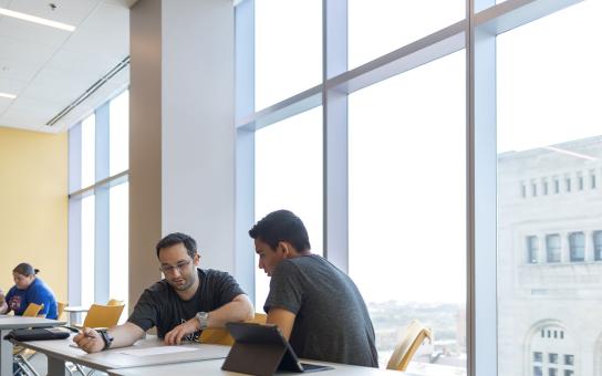 Two students work at a classroom table with the Auditorium tower visible in the windows behind them