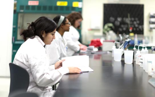 Three students seated a worktable in a lab, the student in the foreground is looking at a book